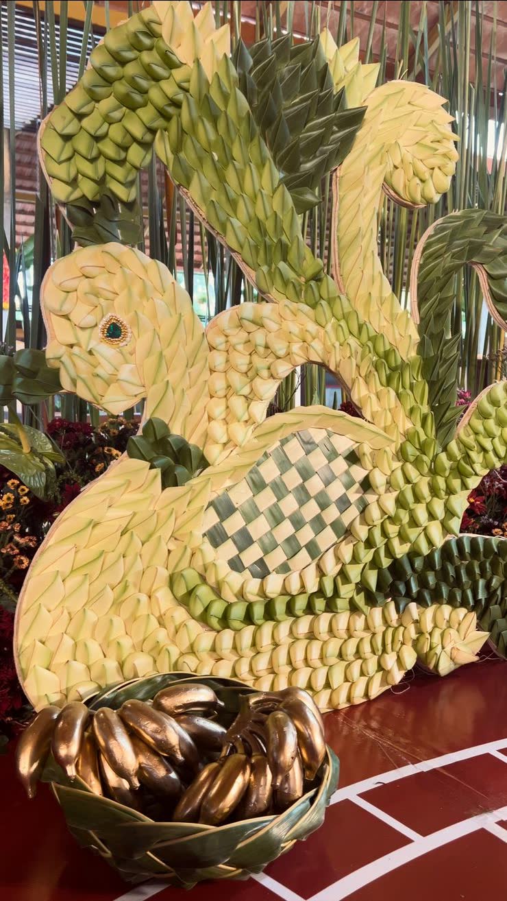 Life-size squirrel sculpture woven from split tender coconut fronds with a green and cream chequer weave panel on the flank and a woven leaf basket of gold-painted bananas at its base