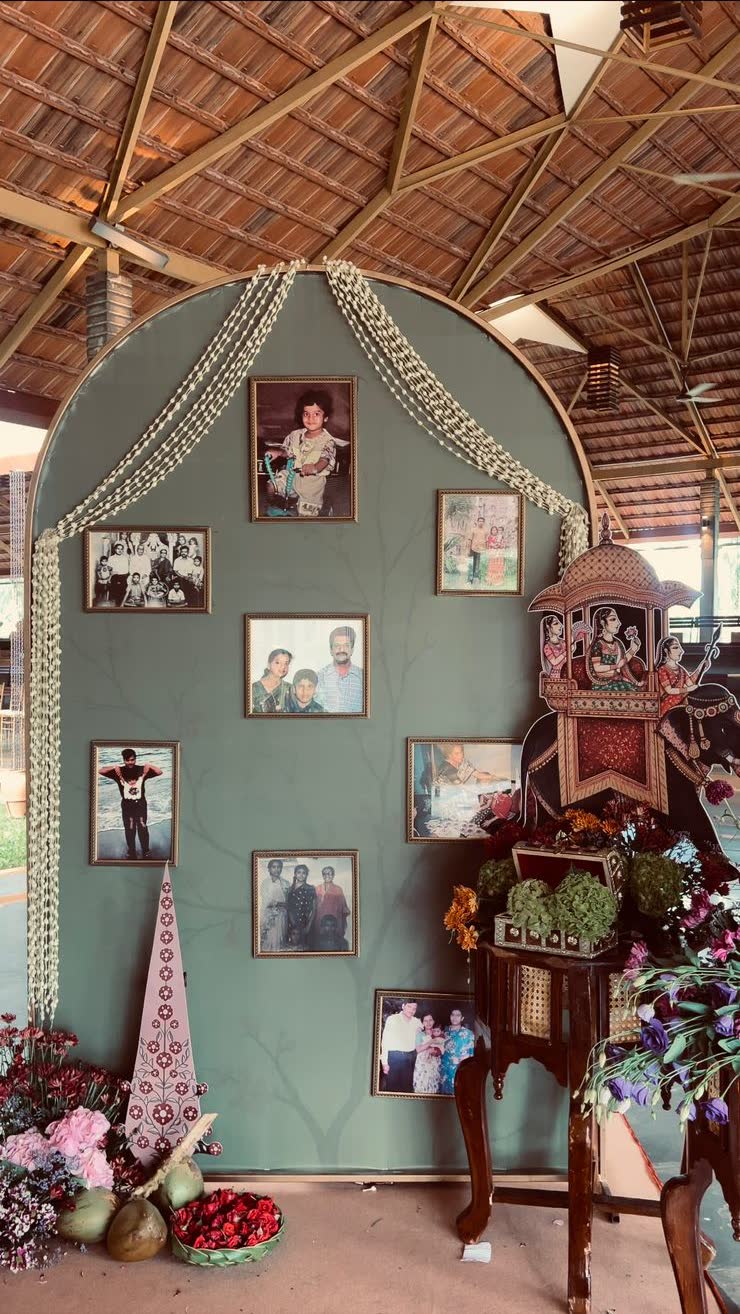 Sage-green painted arch with a bare branch motif hung with the family's framed photographs, jasmine ropes falling from the crown, flowers and coconuts at the base