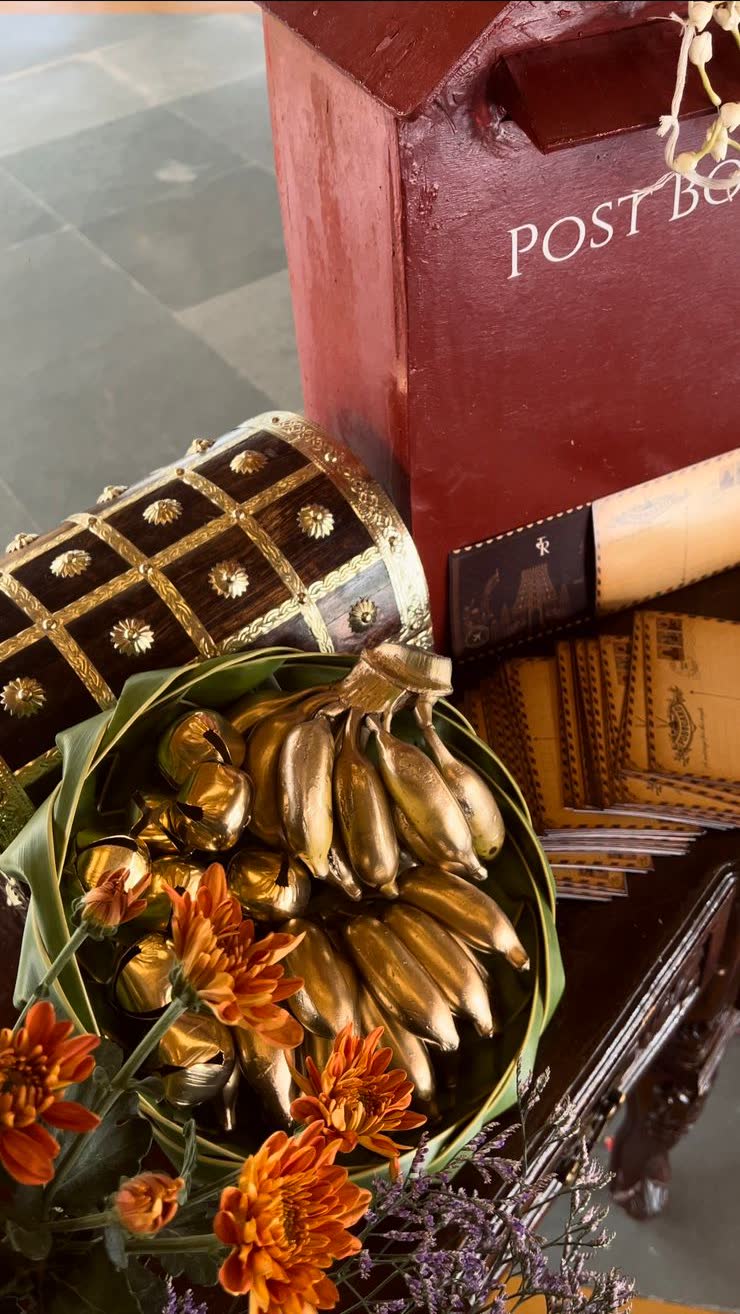 Overhead view of gold-painted bananas arranged on a folded banana leaf plate with orange chrysanthemum, beside an inlaid chest and postcards