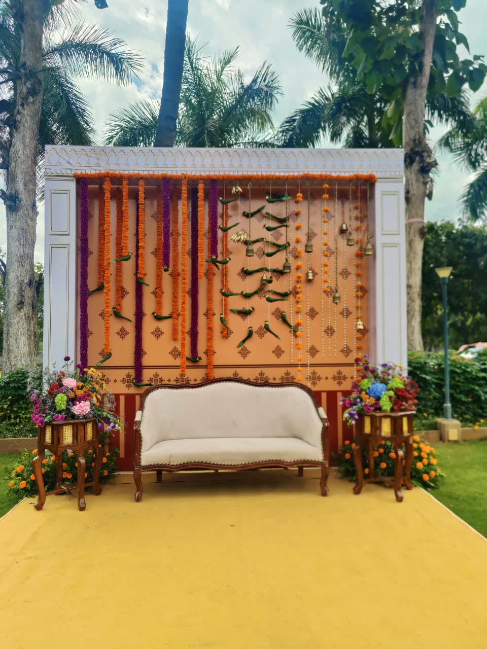 Haldi ceremony backdrop on the lawn — a terracotta panel with printed motif hung with marigold and purple garland ropes, brass bells, green chillies and lemon on thread, a white settee and flower urns
