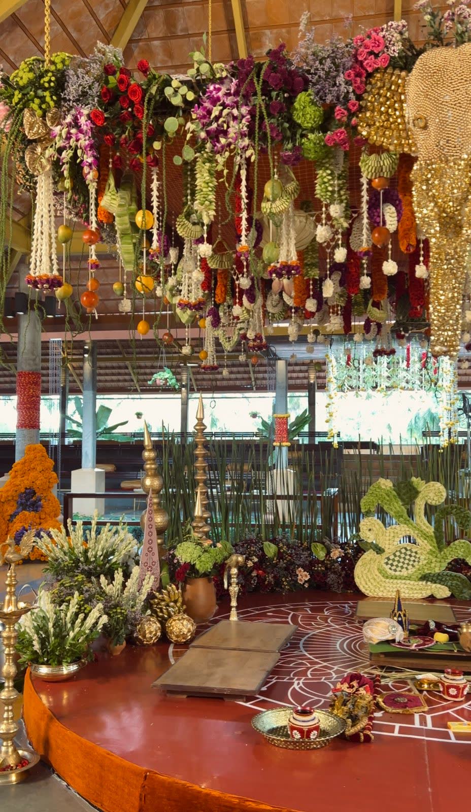 The 360-degree mantap at Panchavati Pavilion with a suspended floral raft overhead, brass finials, a woven coconut-frond squirrel and a red ceremony platform marked with rangoli