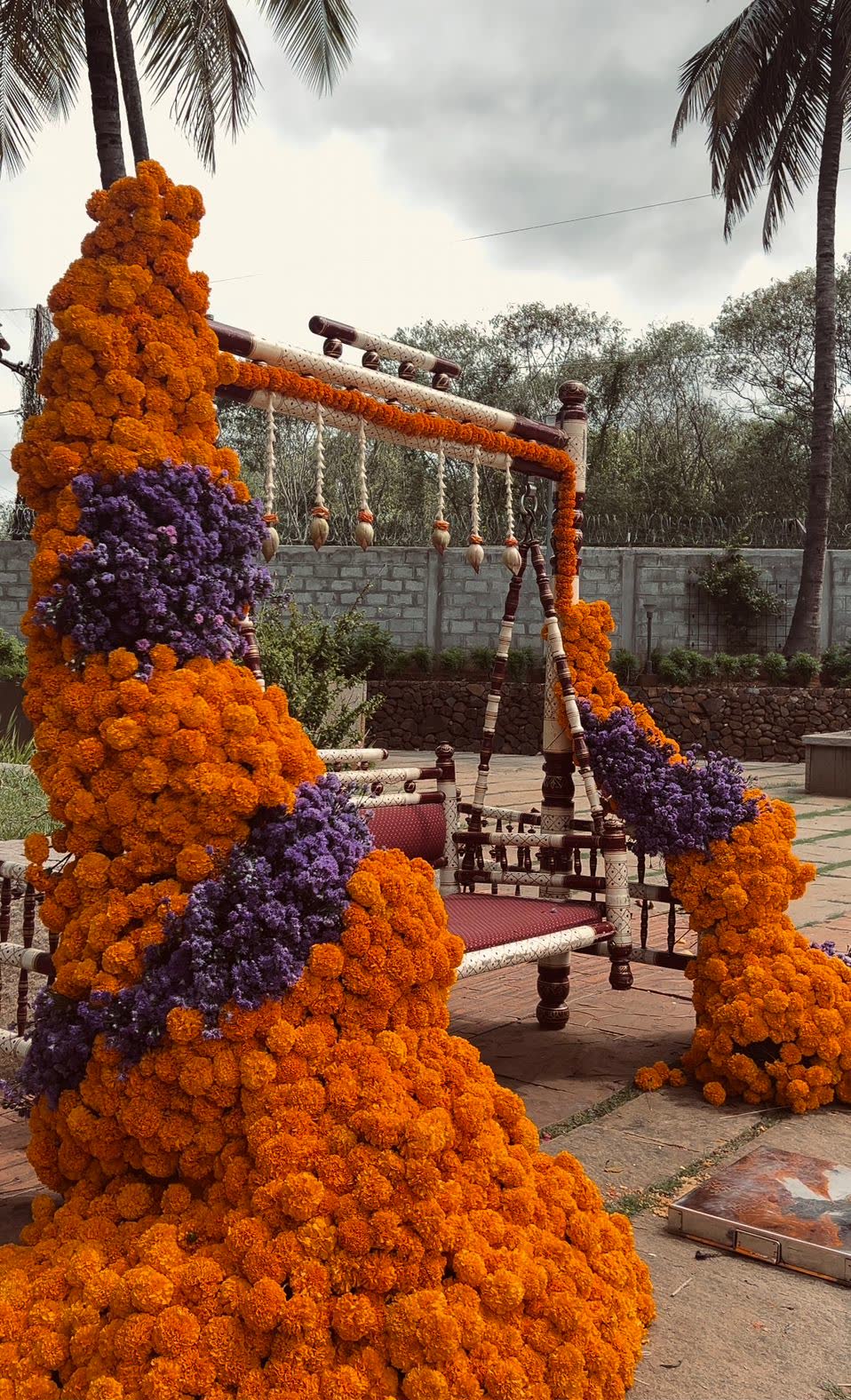 Traditional turned-wood wedding swing banked in orange marigold with purple statice cut through on the diagonal, on the lawn at Panchavati Pavilion