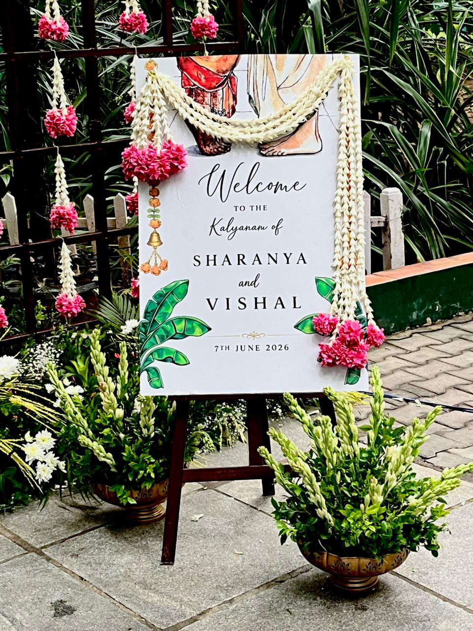 Kalyanam welcome board for Sharanya and Vishal with jasmine garlands and pink kanakambara at MG Magnus Bangalore