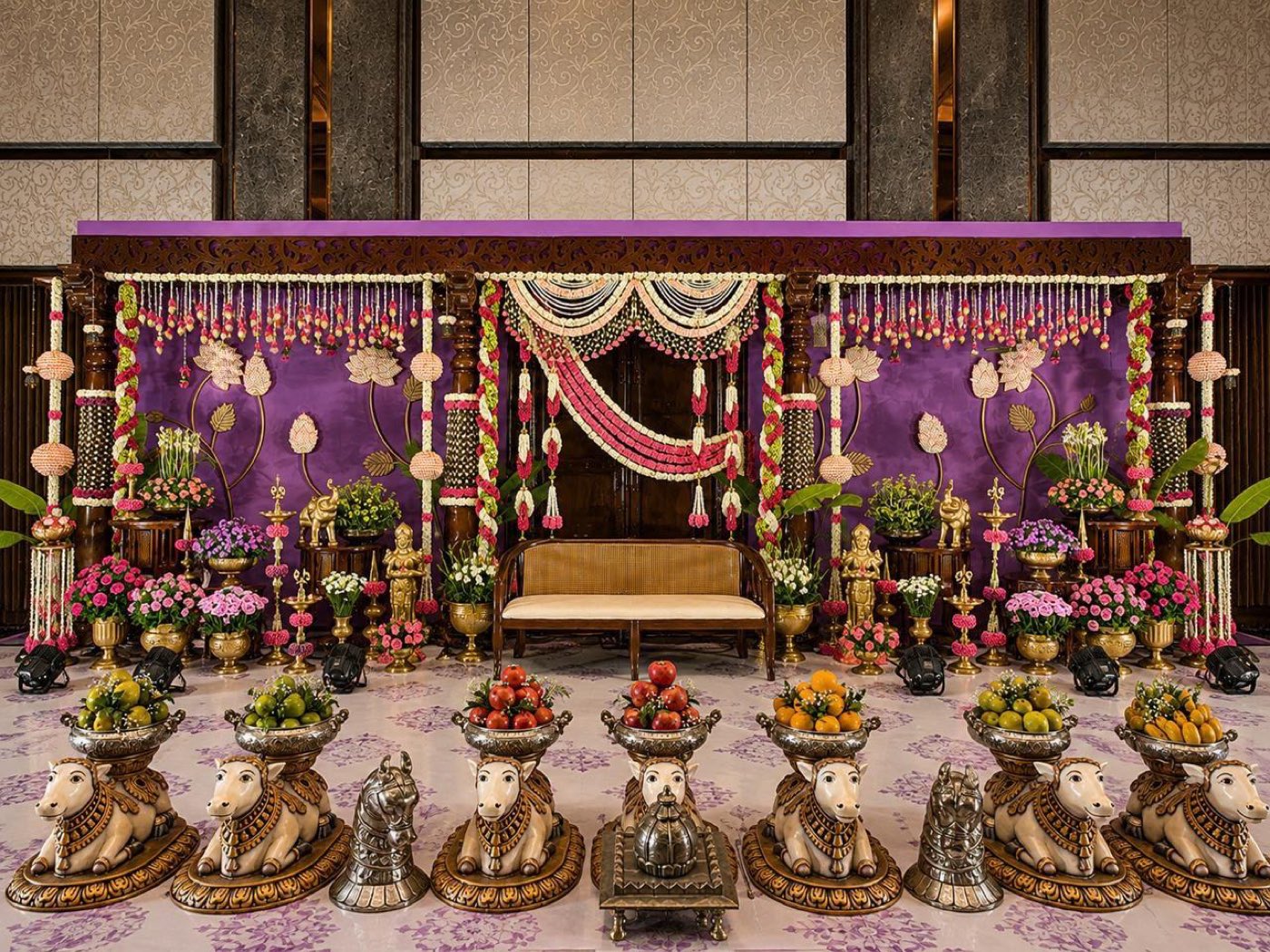 Traditional half saree ceremony stage in purple and pink with carved pillars, jasmine and carnation swags, brass lamps and Nandi figures, at Sheraton Grand Bengaluru Brigade Gateway — designed and built by Panigrahana