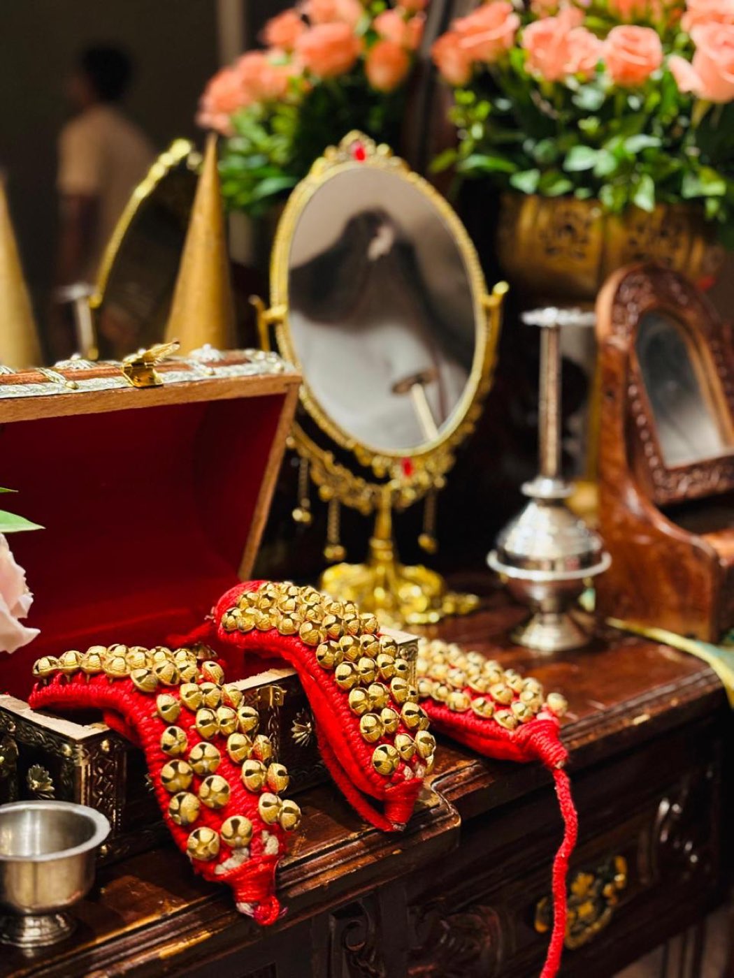 Gejje anklets of gold bells on red cord presented in a carved box beside a brass mirror and peach roses