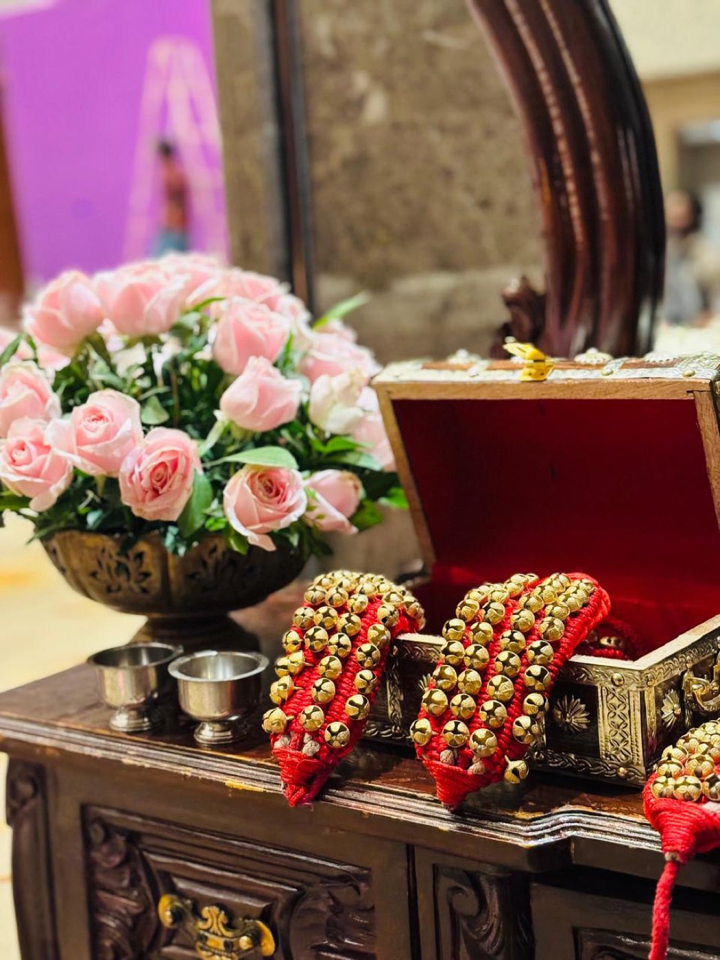 Detail of gejje bell anklets in an open box with a pink rose arrangement and silver ritual vessels