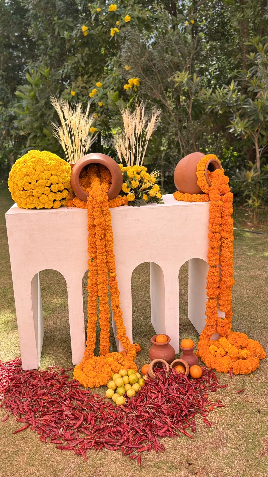 Terracotta pots pouring marigold garlands over a white arched console with wheat, dried chillies, lemons and oranges