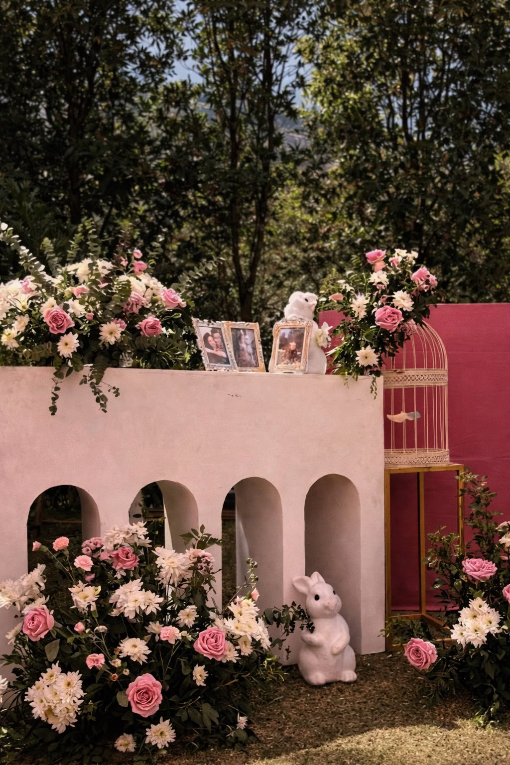 Memory corner with framed photos, birdcage and rabbit figures on a white arched console among pink and white florals