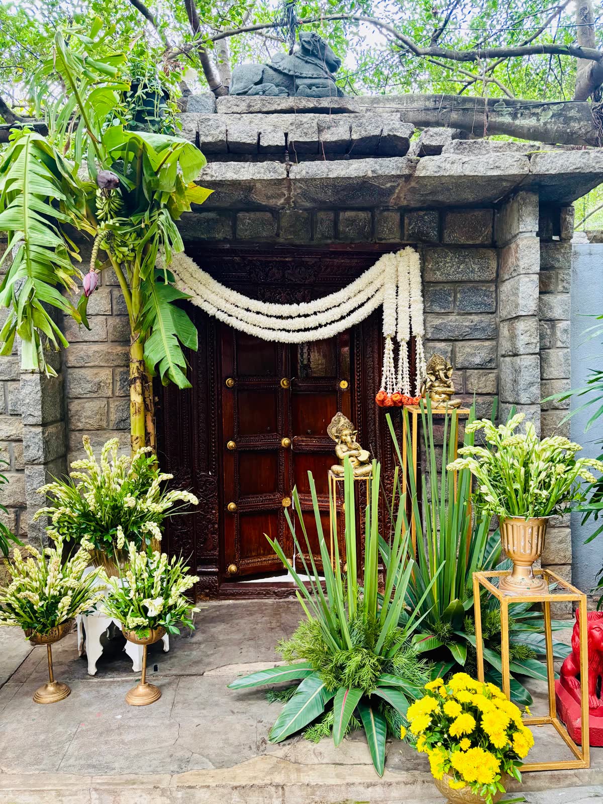 Carved teak double door at The Tamarind Tree with layered white jasmine swags, brass Ganesha figures on stands and urns of tuberose and yellow chrysanthemum
