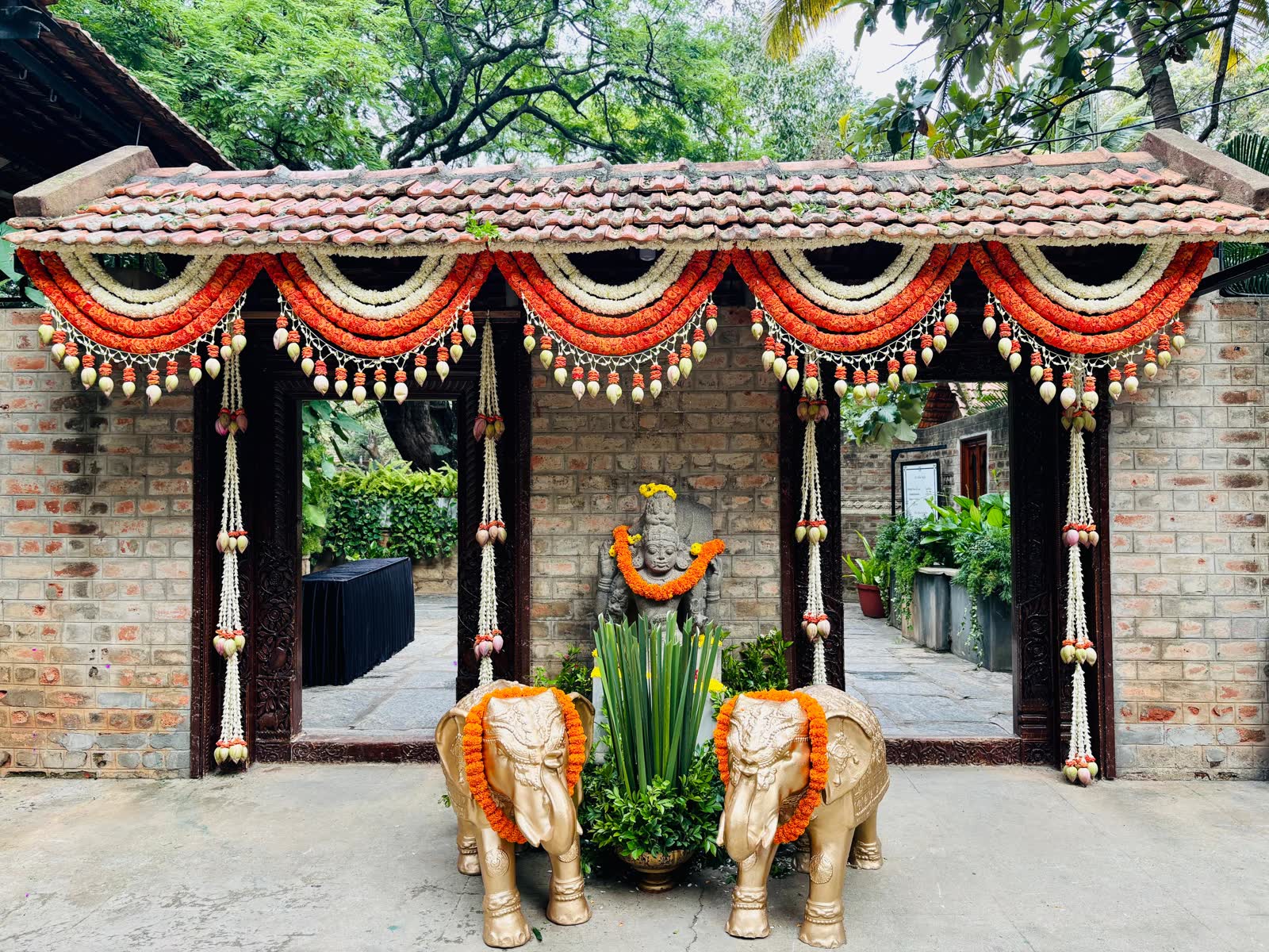 The Vishnu Mantapa portico at The Tamarind Tree dressed with scalloped orange rose and white jasmine swags with lotus bud tassels, two gold elephants garlanded in marigold flanking a stone deity