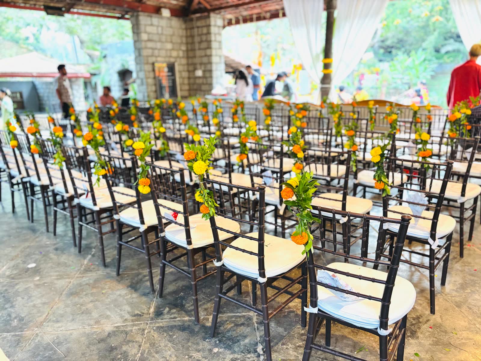 Rows of dark chiavari chairs under the Heritage Pavilion at The Tamarind Tree, each with a single marigold and green foliage sprig wired to the back post