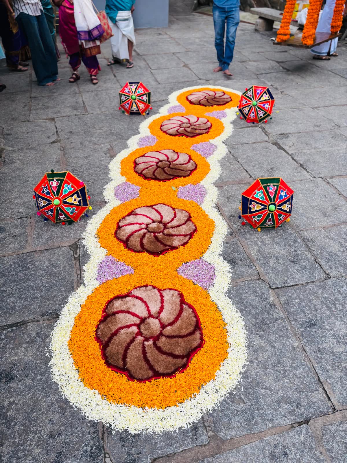 Hand-laid petal aisle runner in white and orange with maroon and cream spiral medallions and purple corner lozenges, flanked by painted kolam umbrellas on granite paving