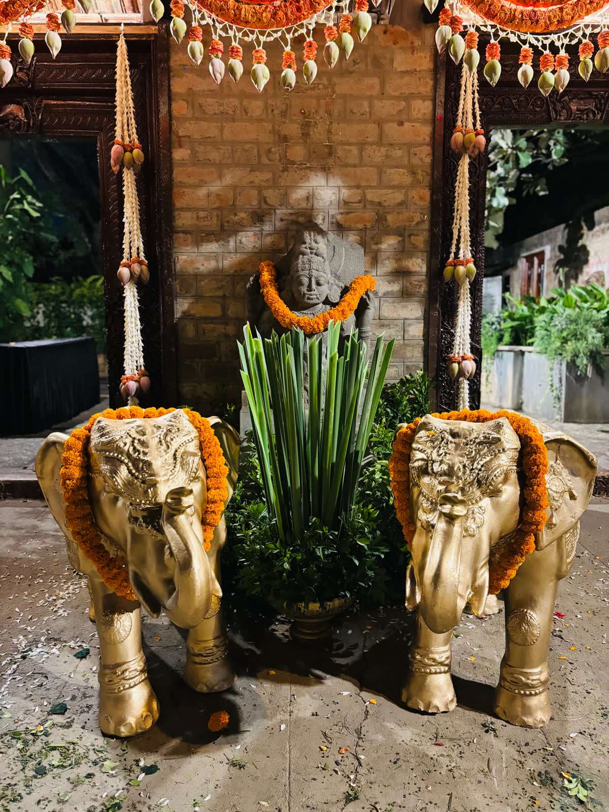 Two gold elephant statues garlanded with marigold flanking a garlanded stone deity in a brick niche, with jasmine and lotus bud tassels hanging above, photographed at night