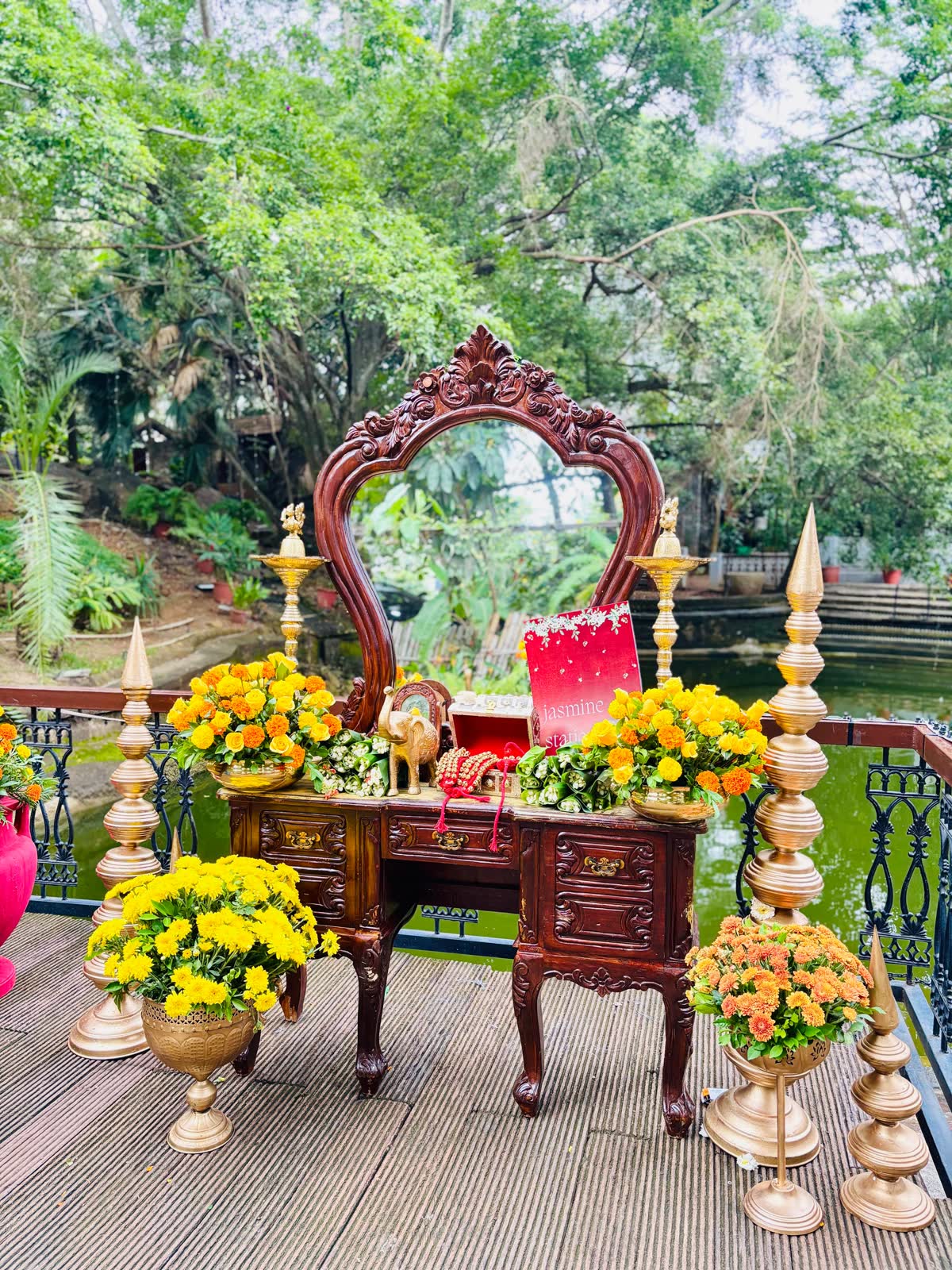 Antique carved wooden vanity with mirror set on the pond deck at The Tamarind Tree, dressed with brass finials, marigold and chrysanthemum urns and trays of strung jasmine