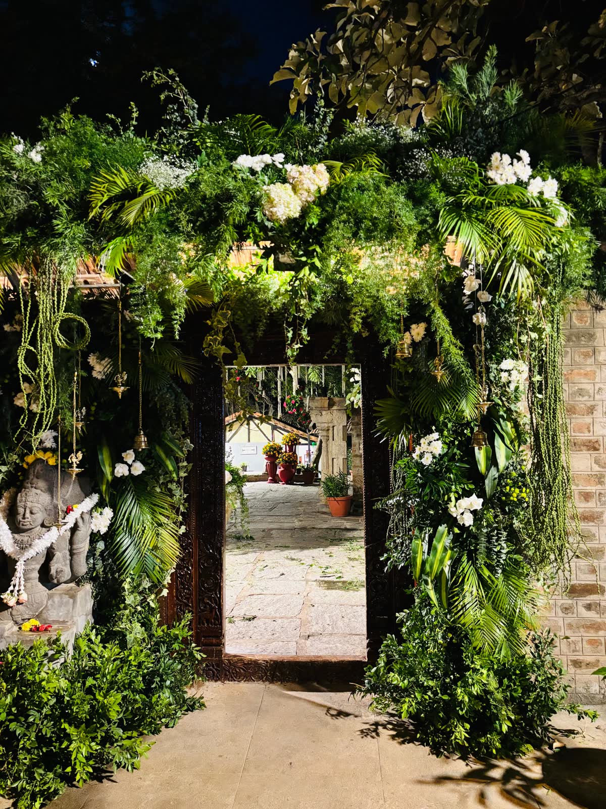 Carved teak doorway at The Tamarind Tree wrapped in dense fern, palm and hanging amaranthus with white orchids, brass bells and hanging oil lamps, lit at night