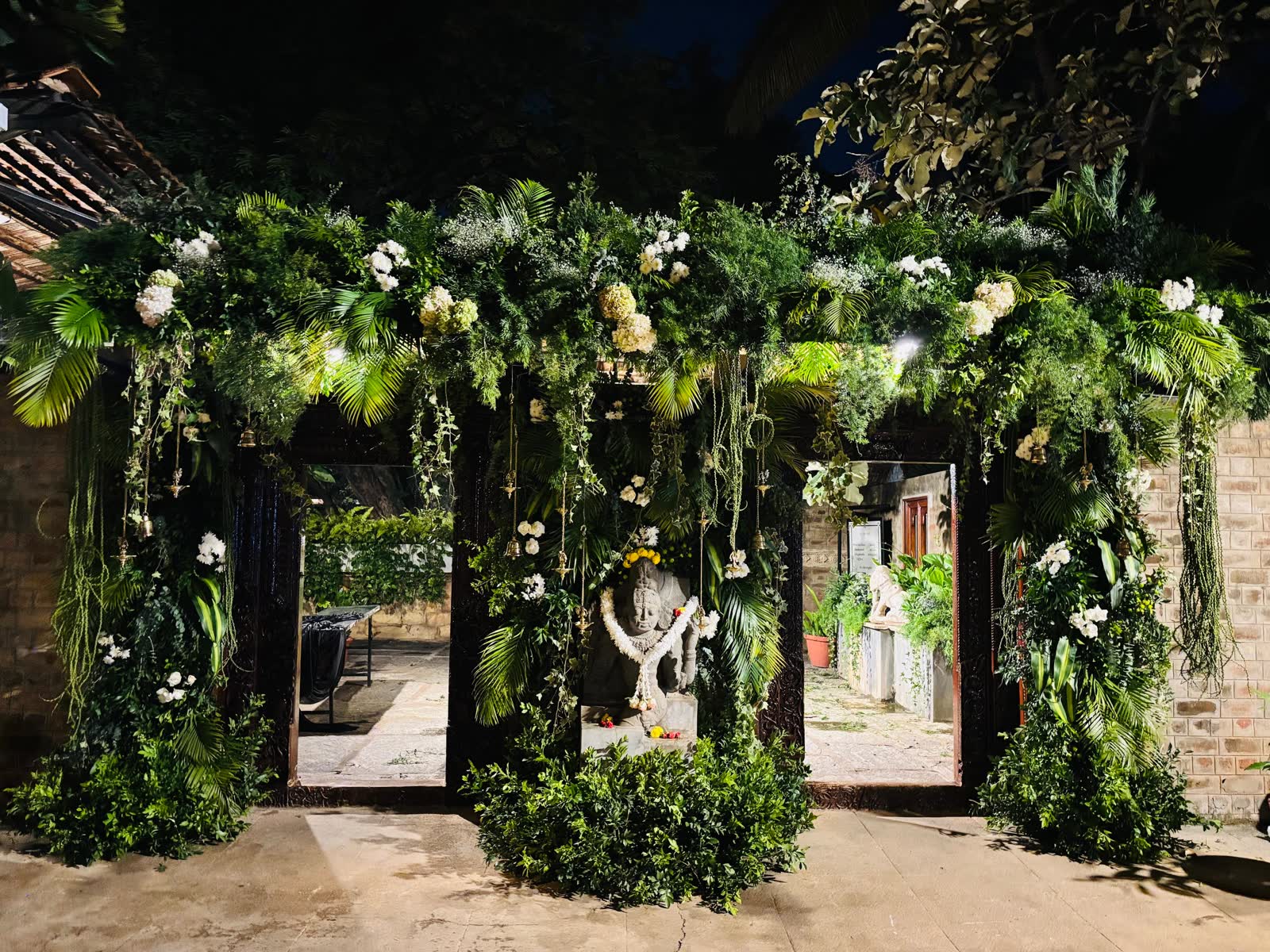 Triple portal at The Tamarind Tree wrapped in dense greenery and white orchids around a garlanded stone deity, lit at night