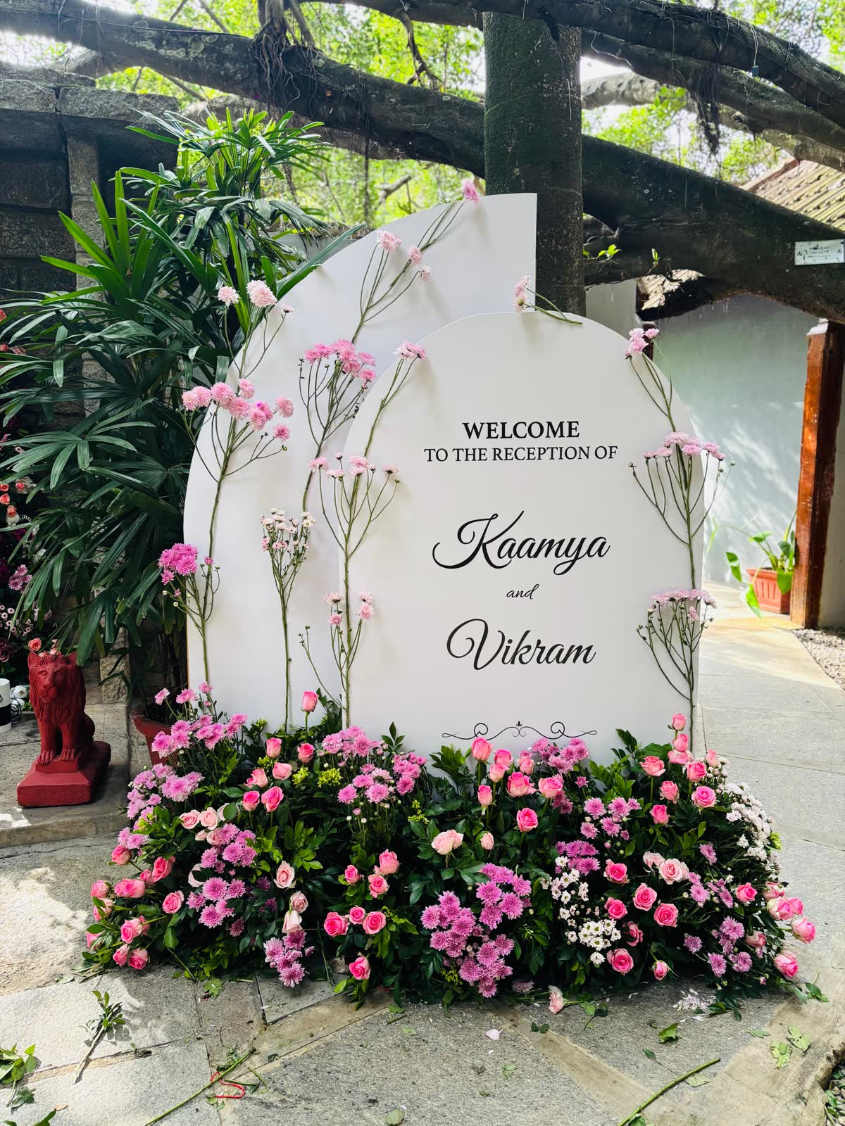 Arched white reception welcome sign for Kaamya and Vikram with tall pink chrysanthemum stems and a dense bank of pink roses and chrysanthemum at its base