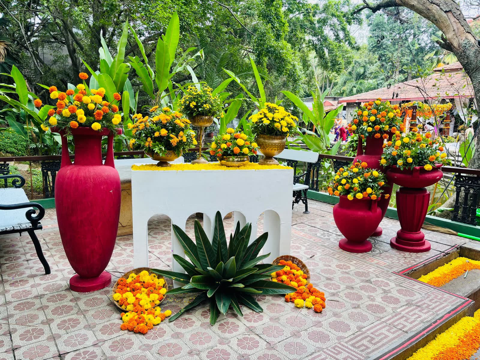 White arched console flanked by tall crimson urns filled with marigold and chrysanthemum, with baskets of loose marigold and a green agave at the base