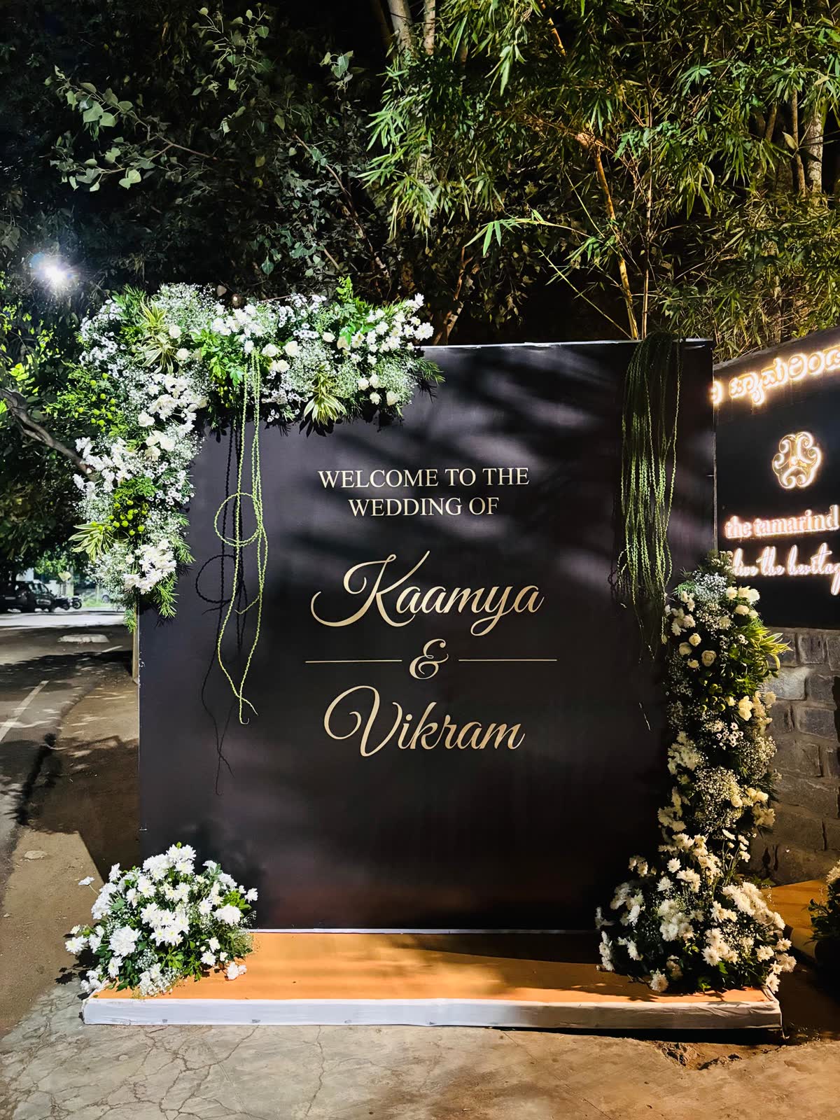 Black welcome board in gold script reading Welcome to the wedding of Kaamya and Vikram, flanked by white flower clusters and green foliage at night at The Tamarind Tree