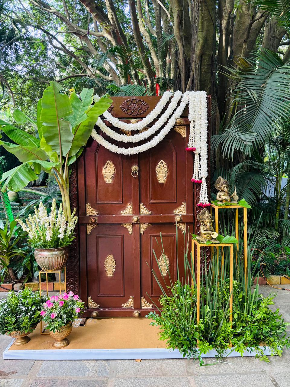 Antique carved door photo backdrop with jasmine swags, banana plant and brass Ganesha idols on gold stands at MG Magnus Bangalore
