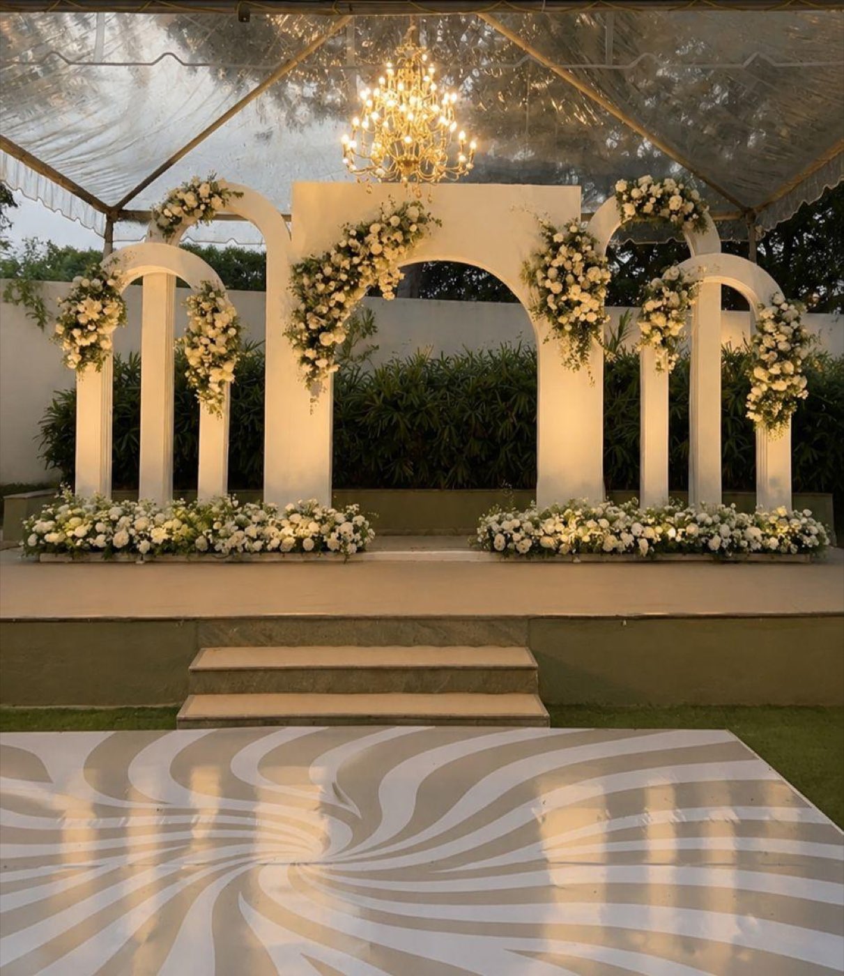 White arched stage with ivory rose and orchid florals under a chandelier, with a patterned dance floor in front, at a 25th wedding anniversary at Farmhouse Social Bangalore — by Panigrahana