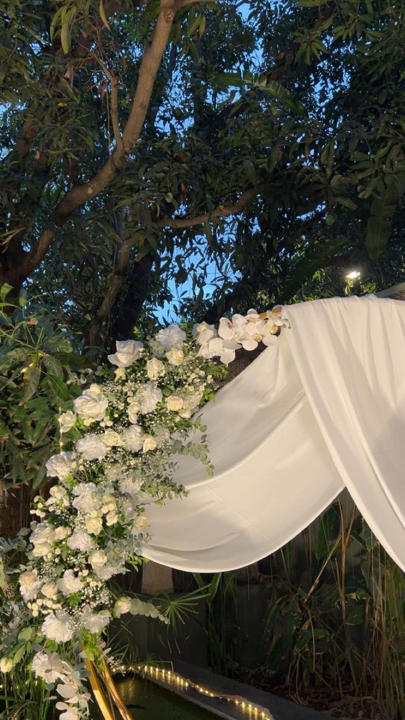 White drape swagged under a tree with an ivory floral cluster at the corner, lit at dusk