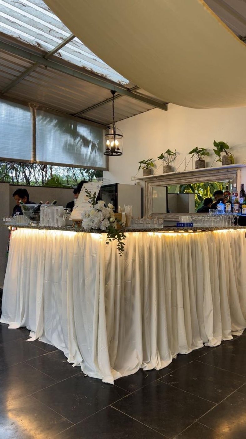 Bar counter fully draped in ivory fabric with warm light under the counter lip and white florals on top, at Farmhouse Social Bangalore
