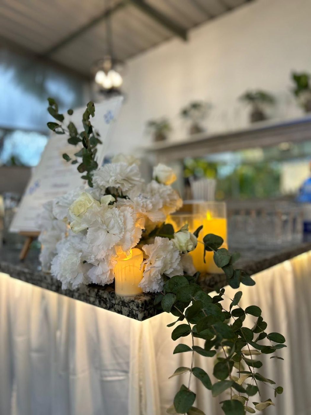 White carnations, roses and candles with trailing eucalyptus arranged on the bar top at an anniversary party